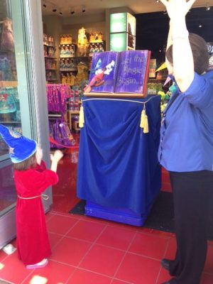 A child dressed as Sorcerer Mickey waves their hands at a magical book, in a store entrance.