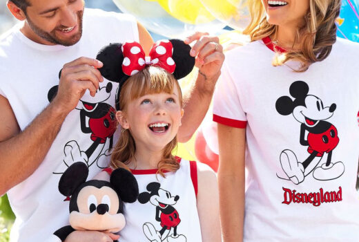 A girl wears Minnie Mouse ears, pinched by her father. Her mother is also in-behind, and they all wear Mickey Mouse t-shirts.