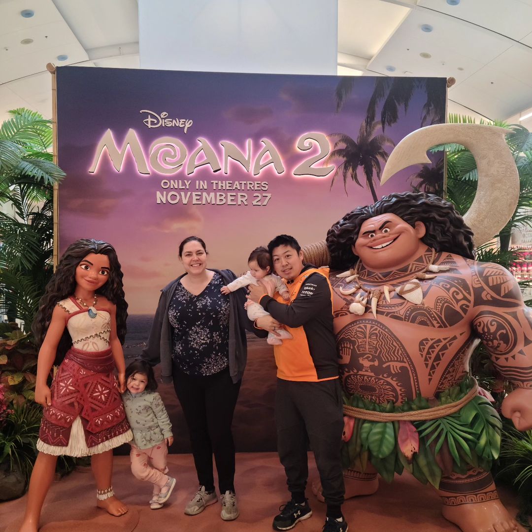 A family poses in front of a backdrop, with statues of Moana and Maui.