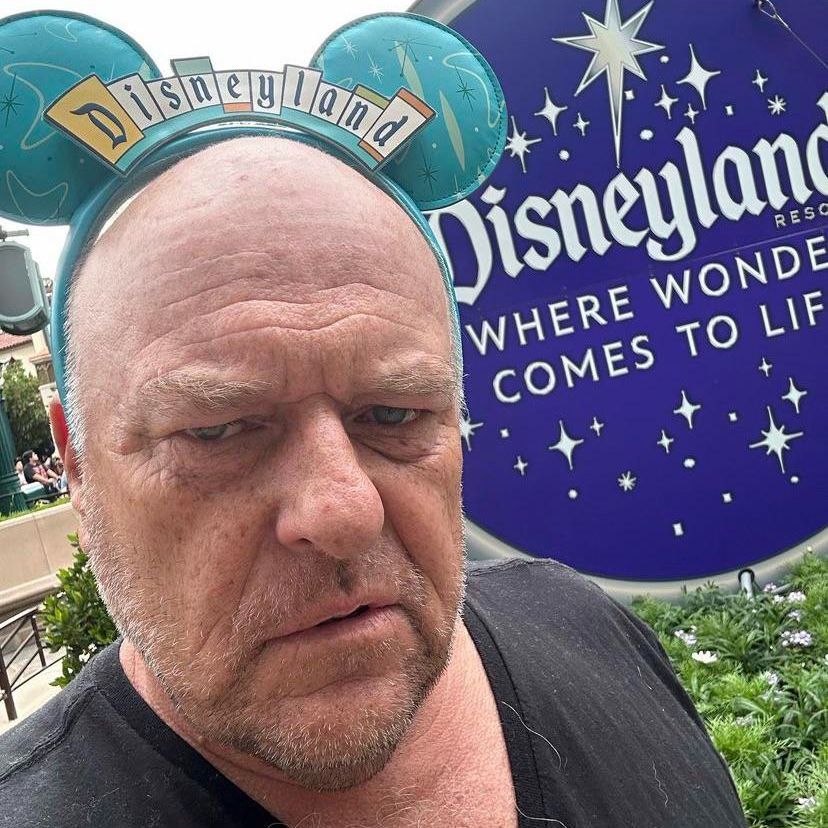 An older, bald man looks grumpy, while wearing Mickey ears with the Disneyland sign.