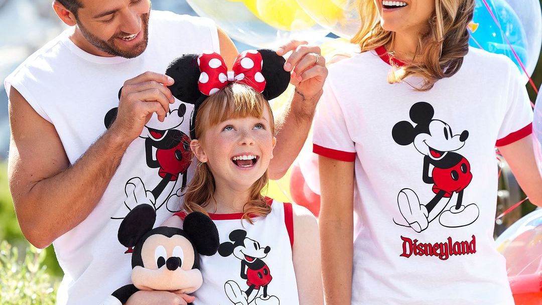 A girl wears Minnie Mouse ears, pinched by her father. Her mother is also in-behind, and they all wear Mickey Mouse t-shirts.