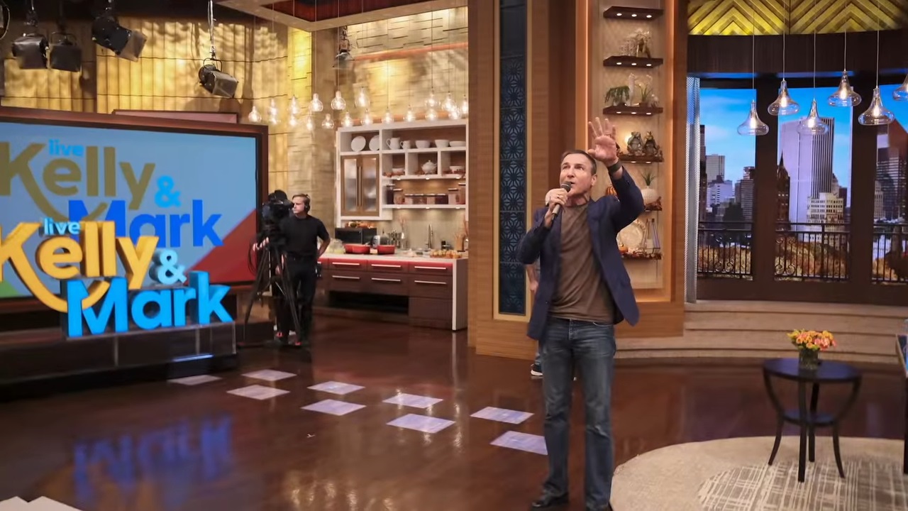 A man raises his hand to an unseen audience behind the camera, on a set.