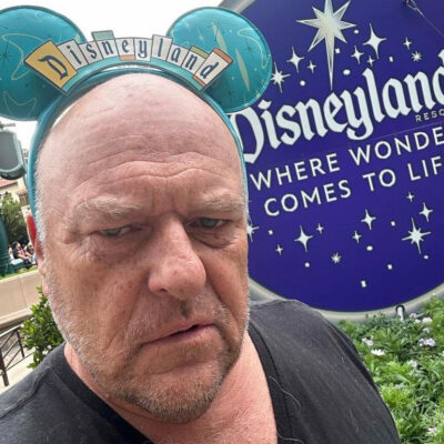 An older, bald man looks grumpy, while wearing Mickey ears with the Disneyland sign.