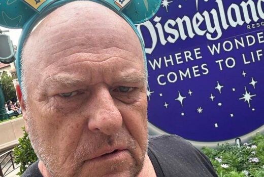 An older, bald man looks grumpy, while wearing Mickey ears with the Disneyland sign.