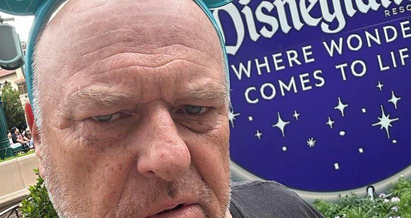 An older, bald man looks grumpy, while wearing Mickey ears with the Disneyland sign.
