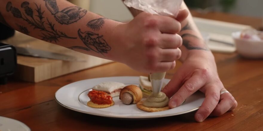 A tattooed arm holds a piping bag, topping a cracker.