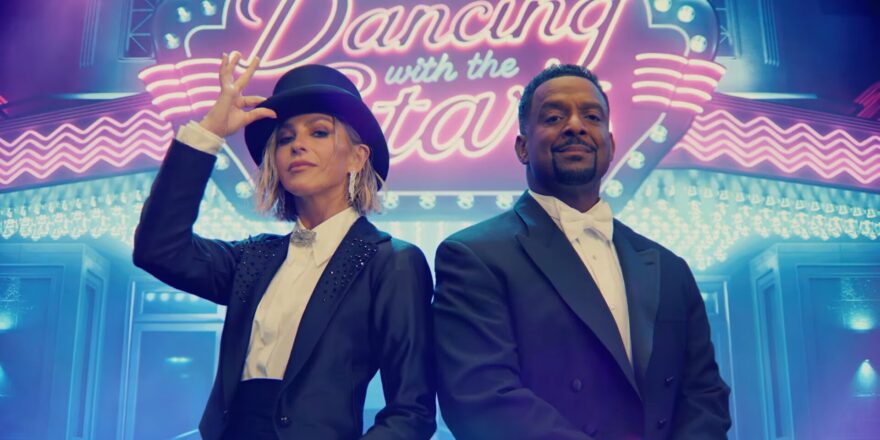 A woman and man in tuxedos, in front of a neon sign.